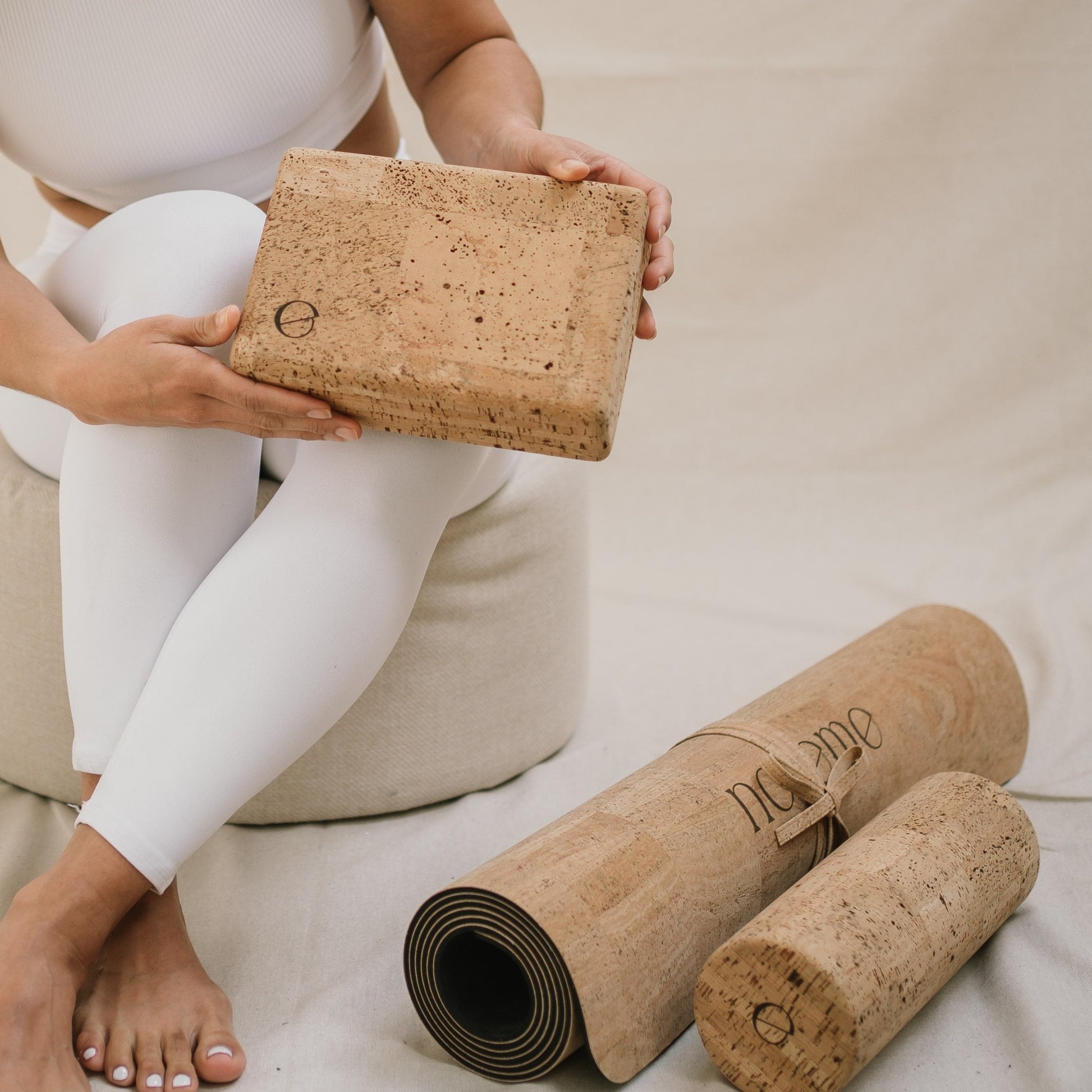 woman holding a Noveme cork yoga block seated next to a Noveme Cork Yoga Mat and Roller